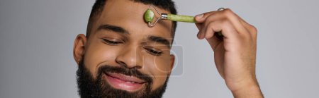 Afrikanisch-amerikanischer junger Mann benutzt Gesichtsroller für Hautpflege-Routine.