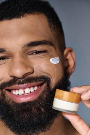 Bearded man joyfully holds jar of cream during skincare routine.