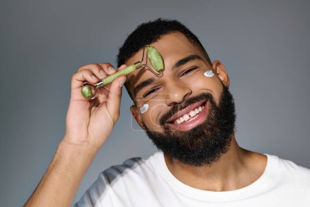 Hombre barbudo afroamericano con crema en la cara usando rodillo facial.