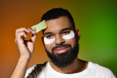 Téléchargez les photos : Un homme avec une barbe et des patchs sur le visage, s'engageant dans une routine de soins de la peau. - en image libre de droit