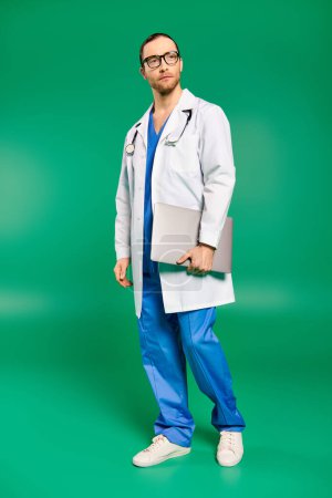 Handsome doctor in white lab coat and blue pants posing against green backdrop.