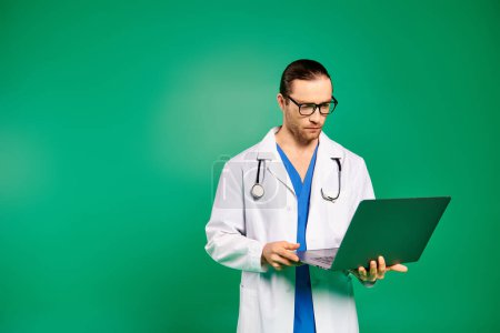 Handsome doctor in white coat holding laptop on green backdrop.