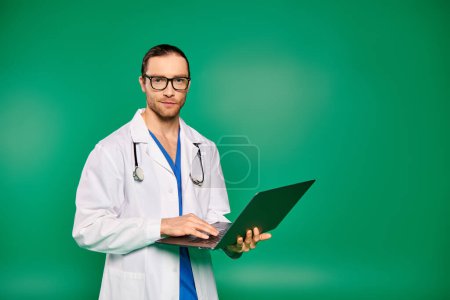 A handsome doctor in a lab coat confidently holds a laptop in front of a green backdrop.