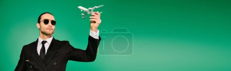 Photo for Handsome pilot in black uniform holding model airplane against green backdrop. - Royalty Free Image