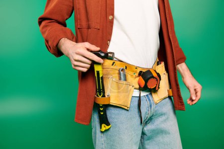 Handsome worker in uniform holding tool belt against green backdrop.