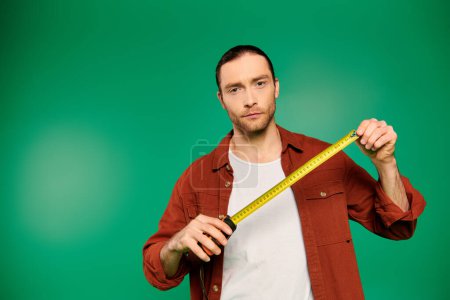 Photo for A handsome male worker in uniform holding a measuring tape against a green backdrop. - Royalty Free Image