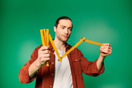 Handsome worker in uniform holds measure tape in front of his face on green backdrop.