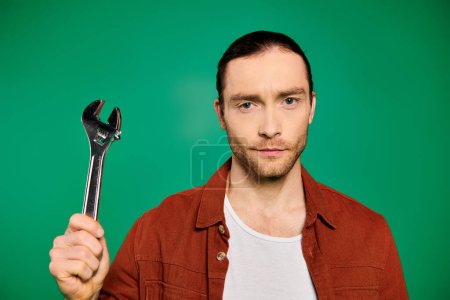 Foto de Un trabajador masculino guapo en uniforme sostiene con confianza una llave inglesa en su mano contra un fondo verde. - Imagen libre de derechos