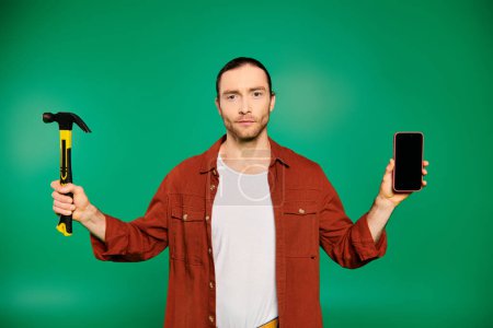 A handsome male worker in uniform holding a hammer and a cell phone on a green backdrop.