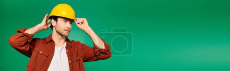 Téléchargez les photos : A handsome male worker in uniform poses with a yellow hard hat on a green backdrop. - en image libre de droit