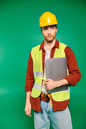A skilled worker in a hard hat confidently holds a laptop in a green backdrop.