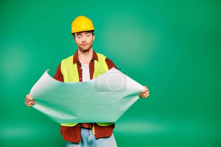 Ein gut aussehender männlicher Arbeiter mit hartem Hut hält ein Papier vor grünem Hintergrund.