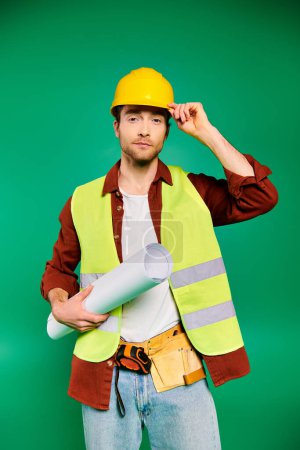 Handsome worker in hard hat and safety vest posing with tools on green backdrop.