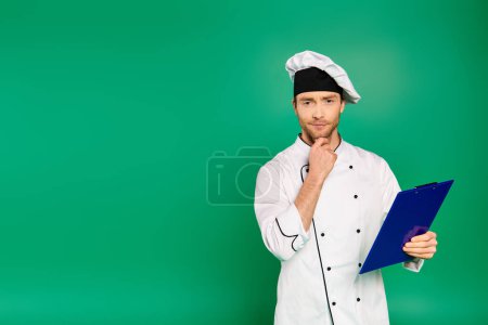 Guapo chef masculino en uniforme blanco sosteniendo un portapapeles sobre fondo verde.