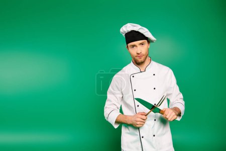 Handsome male chef in white uniform skillfully holding cutlery on green backdrop.