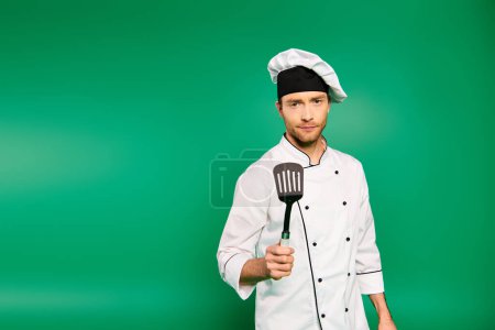 A male chef in a white uniform wields a spatula against a green backdrop.