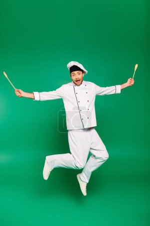 Handsome male chef in white uniform jumping joyfully on green backdrop.