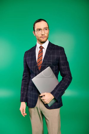 Photo for Elegant businessman in chic suit and tie holding a laptop on green backdrop. - Royalty Free Image