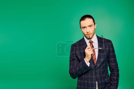 Photo for Handsome businessman in chic suit holding glasses against a green backdrop. - Royalty Free Image