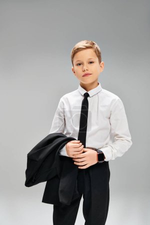 A handsome young boy is dressed in a white shirt and black tie, exuding elegance against a gray backdrop.