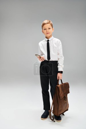 Preadolescent boy in white shirt and tie holds secretive brown bag against gray background.