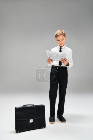 Little boy stands next to suitcase on gray backdrop.