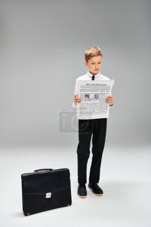 Ein frühpubertärer Junge in eleganter Kleidung steht neben einem Koffer. Geschäftskonzept vor grauem Hintergrund.