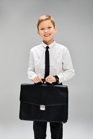 Adorable chico de camisa blanca y corbata sosteniendo un maletín negro sobre un fondo gris.