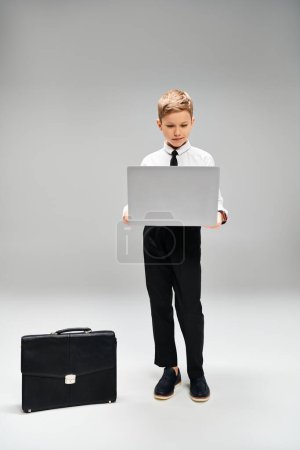 Preadolescent boy in elegant attire holding a laptop next to a suitcase.