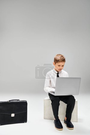 Little boy in smart attire sits on stool absorbed in laptop work, against gray backdrop.
