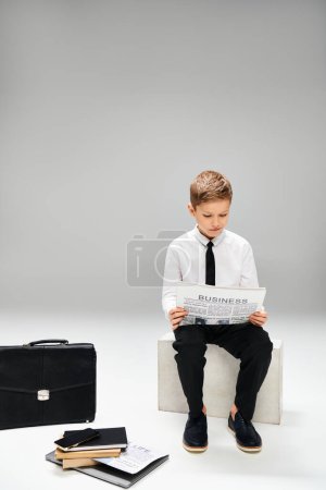Téléchargez les photos : Un garçon préadolescent en tenue élégante s'assoit sur une chaise, absorbé par la lecture d'un journal. - en image libre de droit