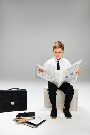 Young boy in elegant attire reading newspaper while seated on stool.