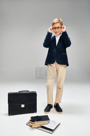 Elegante niño preadolescente de pie con confianza junto a un maletín en un fondo gris.
