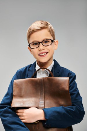 A young boy in elegant attire, wearing glasses, holding a brown bag on a gray backdrop.