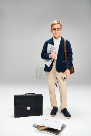 Preadolescent boy in elegant attire stands next to a briefcase against a gray backdrop.