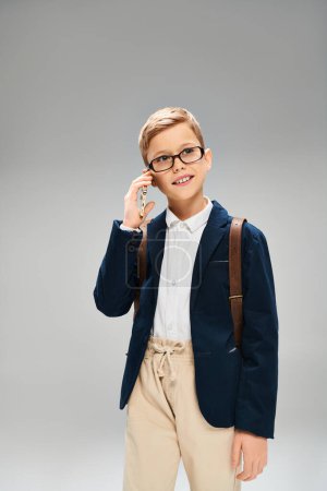 A preadolescent boy with glasses and a jacket standing against a grey backdrop.
