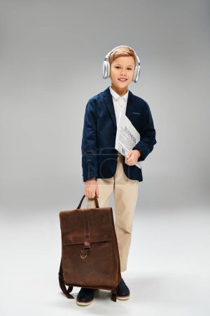 Preadolescent boy in elegant attire, wearing headphones, holds a briefcase on gray backdrop.