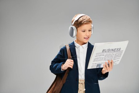 Young boy in elegant attire reads newspaper while wearing headphones.