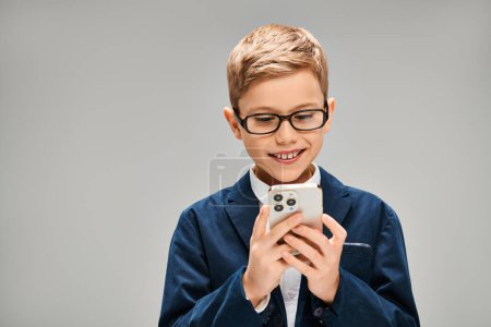 Kleiner Junge mit Brille, Handy in der Hand, elegant gekleidet vor grauem Hintergrund.