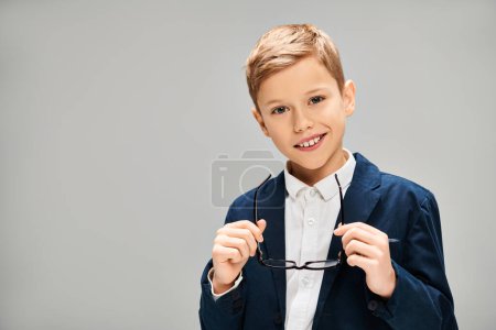 Niño en traje elegante sostiene un par de gafas contra un telón de fondo gris.