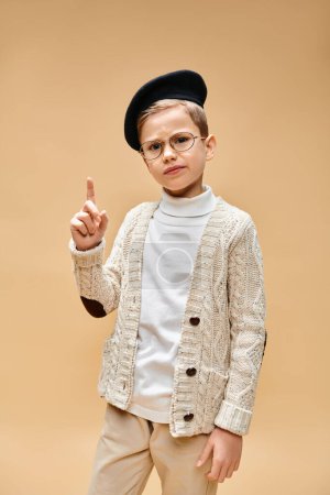 Preadolescent boy in glasses and hat dressed as a film director on beige backdrop.