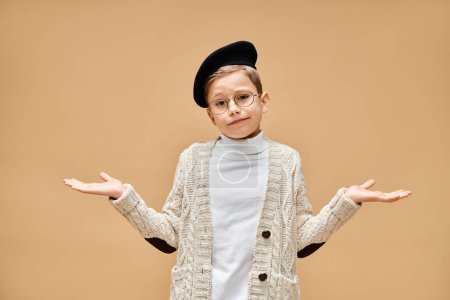 A cute preadolescent boy in glasses and a hat, dressed as a film director on a beige backdrop.