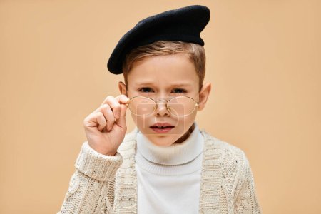 Preadolescent boy in glasses and hat, dressed as a film director on a beige backdrop.