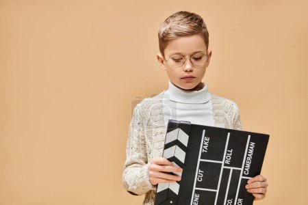 Un lindo niño preadolescente vestido como un director de cine sosteniendo un tablero de aplausos en blanco y negro.