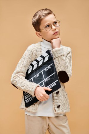 Young boy in glasses holds movie clapboard on beige backdrop.