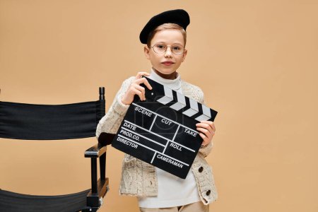 Young boy in film director attire holding a clapper in front of a chair.