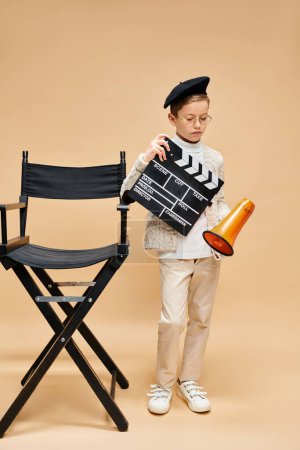 A cute preadolescent boy holding a movie clapper near a chair.
