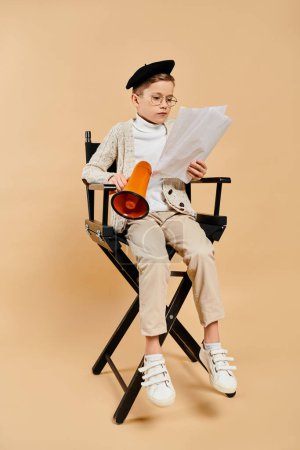 Téléchargez les photos : Un garçon préadolescent, habillé en réalisateur, s'assoit sur une chaise pour lire un journal.. - en image libre de droit