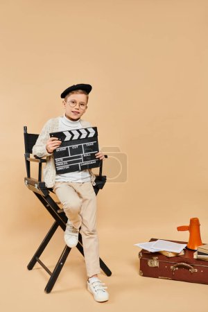 Preadolescent boy in director attire holding movie slate on beige backdrop.
