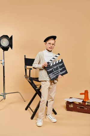 A preadolescent boy in film director attire holds a movie clapper in front of a camera.